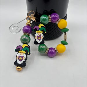 Puede incluir: Un juego de llavero y pulsera con temática de Mardi Gras. El llavero presenta un dije de gnomo y cuentas moradas, verdes y doradas. La pulsera tiene cuentas similares con detalles de pedrería. El fondo incluye un recipiente negro.