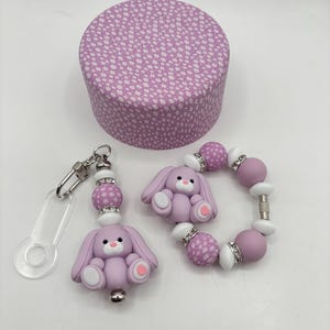 Peut inclure: Un porte-clés, un bracelet et une boîte ronde sur le thème du lapin lavande. Le porte-clés et le bracelet comportent des breloques en forme de lapin et des perles décoratives. La boîte a un motif floral sur le couvercle.