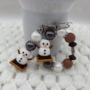May include: A circular keychain featuring two snowman figures sitting on s'mores, interspersed with white, dark gray, and brown beads. The snowman charms have orange noses and black buttons. Silver-toned rhinestone spacers accent the beads.