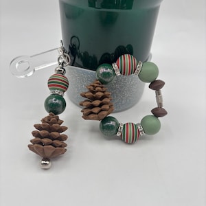 Könnte beinhalten: Ein festliches Armband- und Schlüsselanhänger-Set zum Thema Weihnachten. Das Armband hat grün, rot und weiß gestreifte Perlen sowie grüne und braune Perlen. Der Schlüsselanhänger hat einen Tannenzapfen-Anhänger und passende Perlen. Beide haben silberne Akzente.