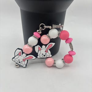 Könnte beinhalten: Ein Schlüsselanhänger mit Perlen und einem Hasenmotiv. Der Schlüsselanhänger hat rosa, weiße und silberne Perlen und einen Hasenanhänger. Der Hasenanhänger ist weiß mit rosa Akzenten. Er ist an einem silbernen Verschluss befestigt.