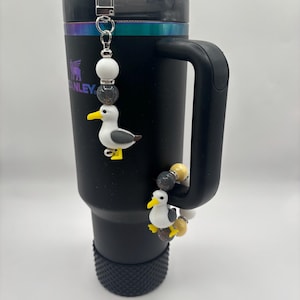 Peut inclure: Gobelet isotherme noir avec une anse, orné d'un charm en perles représentant une figurine de mouette. Le charm comprend des perles blanches, grises et jaunes. Le gobelet a un couvercle noir et une paille argentée.
