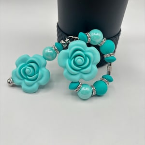 Może przedstawiać: Turkusowy naszyjnik do ząbkowania z dwoma dużymi koralikami w kształcie kwiatów i mniejszymi okrągłymi koralikami. Koraliki są ozdobione srebrnymi dżetami. Naszyjnik jest przeznaczony dla niemowląt i małych dzieci.