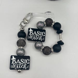Può includere: Un set di portachiavi e braccialetto con perline nere e argentate. Il portachiavi e il braccialetto presentano perline nere, argentate e marroni strutturate. Ogni pezzo ha un ciondolo rettangolare nero con le parole "BASIC WITCH" in bianco. Una silhouette di strega è sopra il testo.