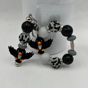 Op de afbeelding: Een sleutelhanger met zwarte glitterkralen, grijze matte kralen en kralen met zwarte vogelsilhouetten. Twee zwarte vogelbedels met oranje snavels zijn bevestigd. De kralen zijn voorzien van zilverkleurige strass-tussenstukjes.