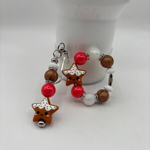 May include: A festive keychain and bracelet set featuring star-shaped gingerbread men with smiling faces and white icing details. The keychain has red, white, and brown beads. The bracelet has red, white, and gold beads. A fun holiday accessory.