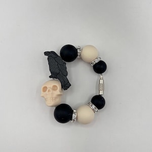 Op de afbeelding: Een zwart en crèmekleurig kralenarmband met een raven- en schedelbedel. De armband heeft zwarte en crèmekleurige ronde kralen, geaccentueerd met zilverkleurige strass-afstandhouders. Een zwarte raaf en een crèmekleurige schedel zijn aan de armband bevestigd.