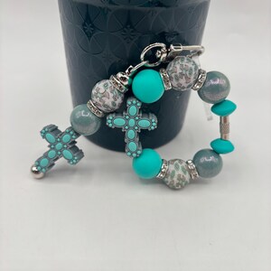 May include: A beaded keychain with a cross design. The keychain features turquoise, grey, and patterned beads with silver accents. The cross is turquoise with a grey border. The keychain is attached to a silver clasp.