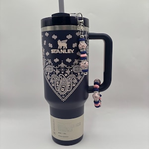 Puede incluir: Un vaso Stanley H2.0 de 1,18L azul marino con un diseño de cachemira blanco y una pajita plateada. El vaso tiene un asa y un charm de cuentas rosas, azules y plateadas. El vaso tiene el logotipo de Stanley.
