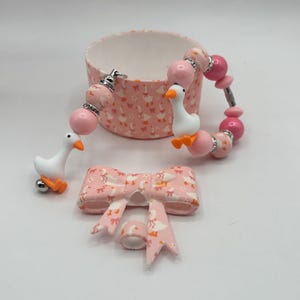 May include: A pink and white beaded bracelet with goose charms, a matching bow, and a small round container. The bracelet features pink beads and goose charms with orange beaks and feet. The bow and container have a pink goose pattern.