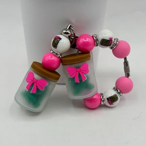 Op de afbeelding: Een sleutelhanger met twee doorzichtige potjes gevuld met groen materiaal en een roze strik. De sleutelhanger heeft ook roze en witte kralen met zilveren accenten. De potjes hebben bruine deksels.