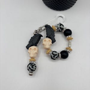Op de afbeelding: Een sleutelhanger- en armbandset met een gotisch thema. De sleutelhanger heeft een zwarte raaf en een crèmekleurige schedelbedel, samen met een zwart-wit patroonkraal. De armband heeft zwarte kralen, patroonkralen en schedelbedels.