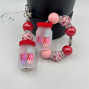 Pode incluir: Um porta-chaves de contas com detalhes vermelhos, rosa e prateados. O porta-chaves apresenta dois pequenos frascos foscos com tampas vermelhas, cada um com o texto "XOXO". O design inclui contas vermelhas e rosa e um fecho prateado.