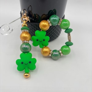 May include: A gold-toned keychain and bracelet set featuring green shamrock charms and beads. The keychain has a clear plastic attachment. The bracelet and keychain have gold and green beads with rhinestone accents. The shamrocks have smiling faces.