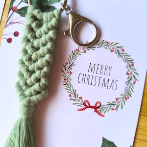 Pode incluir: Um porta-chaves de macramé verde feito à mão com um fecho prateado, combinado com um cartão de Natal. O cartão apresenta uma coroa de aquarela com bagas vermelhas e o texto "FELIZ NATAL".