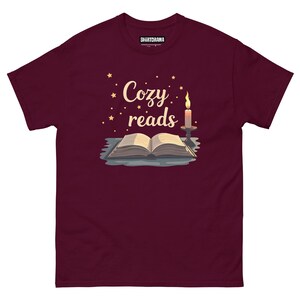 Könnte beinhalten: Burgunderfarbenes T-Shirt mit dem Text "Cozy reads" in goldenen Buchstaben. Das Design zeigt ein Buch mit einer brennenden Kerze und Sternen.
