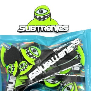 Peut inclure: Un paquet d'accessoires de la marque Subtronics. Les accessoires présentent un motif d'œil de monstre vert fluo avec le mot "Subtronics" en blanc. Les accessoires sont noirs et vert fluo. L'emballage est bleu clair.