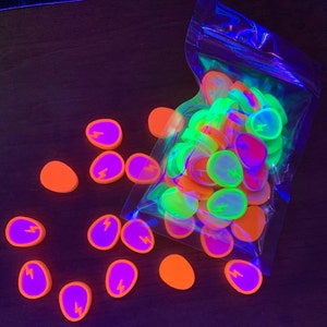 Op de afbeelding: Een zak met neonoranje, groene en paarse platte hars cabochons met een bliksemontlading ontwerp. De cabochons zitten in een doorzichtige plastic zak en zijn verspreid over een donkerbruin oppervlak.