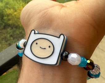 Könnte beinhalten: Ein Perlenarmband mit einem zentralen Anhänger in Form eines Charakters in Weiß, Schwarz und Beige mit lächelndem Gesicht. Das Armband ist mit verschiedenen runden Perlen in durchscheinendem Weiß, leuchtendem Blau und schillerndem Schwarz besetzt.