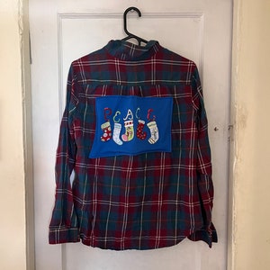 Peut inclure: Une chemise à carreaux à manches longues rouge, verte et bleu marine, suspendue à un cintre noir. Un patch bleu au dos porte le mot "PEACE" et des bas de Noël brodés.