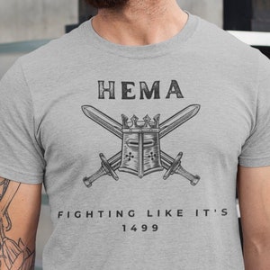Puede incluir: Una camiseta gris con un gráfico negro de dos espadas cruzadas sobre un casco con una corona. El texto "HEMA FIGHTING LIKE IT'S 1499" está impreso debajo del gráfico.