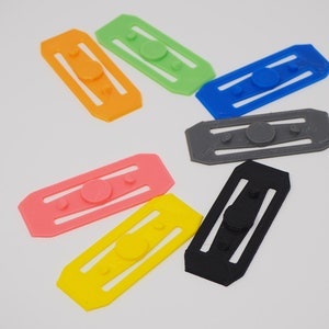 May include: Seven colourful plastic finger fidget toys. Each toy has a round button in the centre and two parallel lines on either side. The colours are green, orange, blue, grey, pink, yellow, and black.