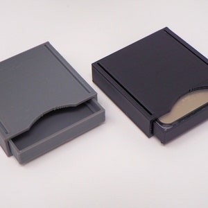 May include: Two small, square storage boxes with lids. One box is grey and the other is black. Both boxes are open and have a small, rectangular object inside.