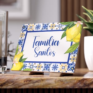 Könnte beinhalten: Ein rechteckiges Schild mit den Worten "Familia Santos" in blauer Schrift. Das Schild hat einen blauen Rand mit floralen und geometrischen Mustern sowie Illustrationen von Zitronen und Blättern. Es wird von zwei Holzstützen gehalten.