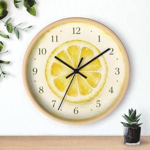 Puede incluir: Un reloj de pared redondo con marco de madera clara. La esfera del reloj presenta una ilustración de acuarela de una rodaja de limón. Las manecillas negras indican la hora. Los números son pequeños y negros, sobre un fondo crema.