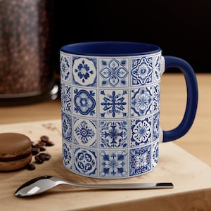May include: A white ceramic mug with a blue and white patterned design. The mug has a blue rim and handle. The pattern is made up of various geometric shapes and floral motifs.