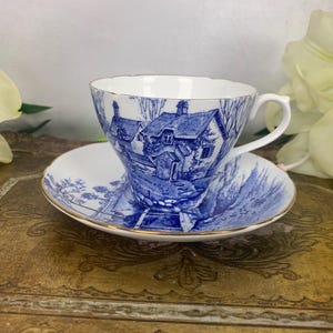 Puede incluir: Un juego de taza y platillo de té de porcelana azul y blanco con una escena de cabaña. La taza y el platillo tienen un borde dorado.