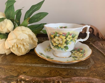 May include: A white teacup and saucer with a floral pattern featuring yellow primroses. The cup and saucer have a gold trim.