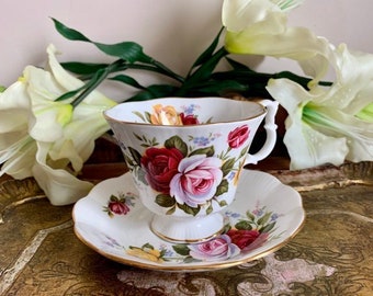 May include: A white teacup and saucer with a floral pattern featuring pink, red, and yellow roses. The cup has a gold rim and a delicate handle. The saucer has a matching floral pattern and a gold rim.