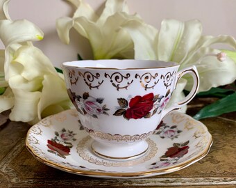 Pode incluir: Uma chávena de chá e um pires brancos com uma borda dourada e um padrão floral com rosas vermelhas e cor-de-rosa.