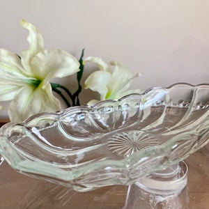 May include: A clear glass serving dish with a scalloped edge and a starburst design in the centre. The dish is oval-shaped and sits atop a clear glass base. White lilies are in the background.