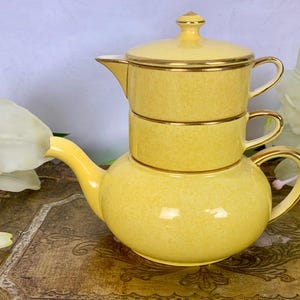 Op de afbeelding: Een gele theepot van keramiek met een gouden rand. De theepot heeft drie verdiepingen en staat op een grotere basis. De theepot staat op een houten oppervlak.