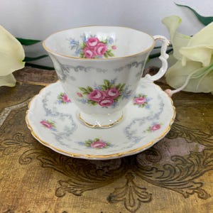 Puede incluir: Un juego de taza y platillo de té blanco con un delicado patrón floral con rosas rosas y detalles azules. El juego tiene un borde dorado.