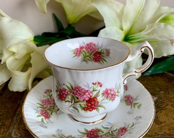 May include: A white teacup and saucer with a floral pattern. The pattern features pink and red flowers with green leaves on a white background. The cup and saucer have a gold trim.