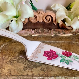 Puede incluir: Un servidor de pastel de cerámica blanca con un borde dorado y un diseño floral con rosas rojas. El servidor está en una bandeja dorada.
