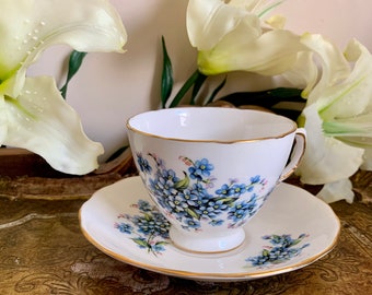 May include: A white teacup and saucer with a blue forget-me-not floral pattern. The cup and saucer have a gold trim.