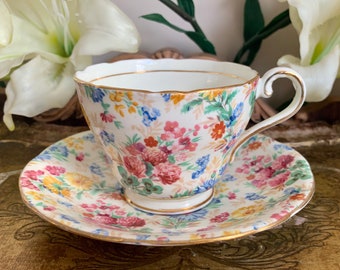 May include: A vintage teacup and saucer set with a floral pattern in shades of pink, blue, yellow, and green. The set has a gold trim around the edges.