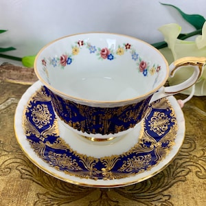 Könnte beinhalten: Eine weiße Teetasse und Untertasse mit einem blauen und goldenen Blumenmuster. Die Tasse hat einen zarten Blumenschmuck am Rand und die Untertasse ein filigraneres Muster.