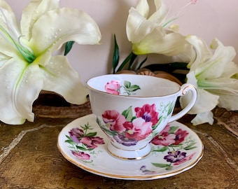 May include: A white teacup and saucer with a floral pattern of pink, purple, and green flowers. The cup and saucer have a gold trim.