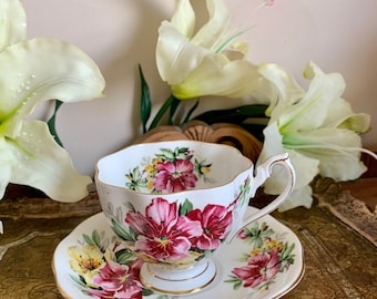 Puede incluir: Una taza de té y un platillo blancos con un patrón floral con flores rosas, amarillas y verdes. La taza y el platillo tienen un borde dorado.
