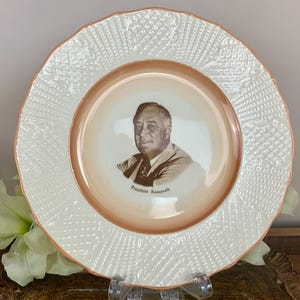 May include: A white ceramic plate with a raised, textured design around the edge. The centre of the plate features a brown image of President Roosevelt.