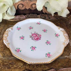 May include: A white porcelain serving tray with a gold rim and handles. The tray is decorated with a floral pattern of pink roses and green leaves.