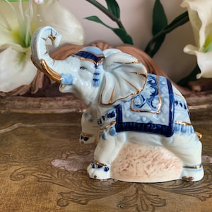 May include: A white porcelain elephant figurine with blue and gold accents. The elephant is standing on a base and has its trunk raised. The figurine is decorated with a blue and gold patterned saddle.