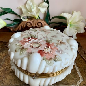 May include: A white, round, hinged trinket box with a floral design. The box has a raised, textured rim and a brass hinge.