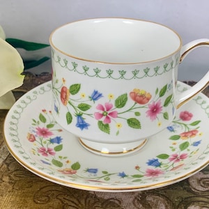 Op de afbeelding: Een wit theekopje en schoteltje met een bloemenpatroon in tinten roze, blauw, geel en groen. Het kopje en schoteltje hebben een gouden rand.
