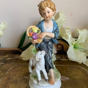 May include: A porcelain figurine of a boy holding a basket of fruit, standing next to a white lamb. The boy is wearing a blue jacket and black pants. The figurine is on a white base.